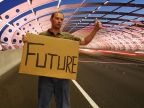 Ein Mann will als Anhalter mitgenommen werden, auf einem Pappschild hat er sein Ziel angegeben: Future - Zukunft.